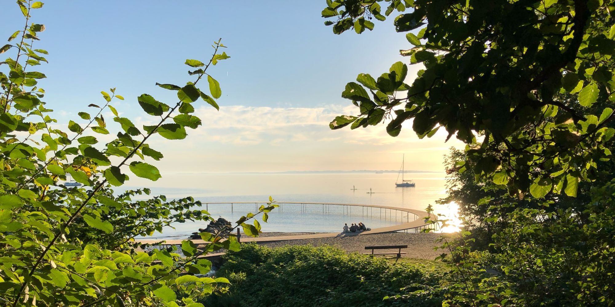 Landscape of Danish beach and sea at sunrise, with surfers and sailboat in the background.